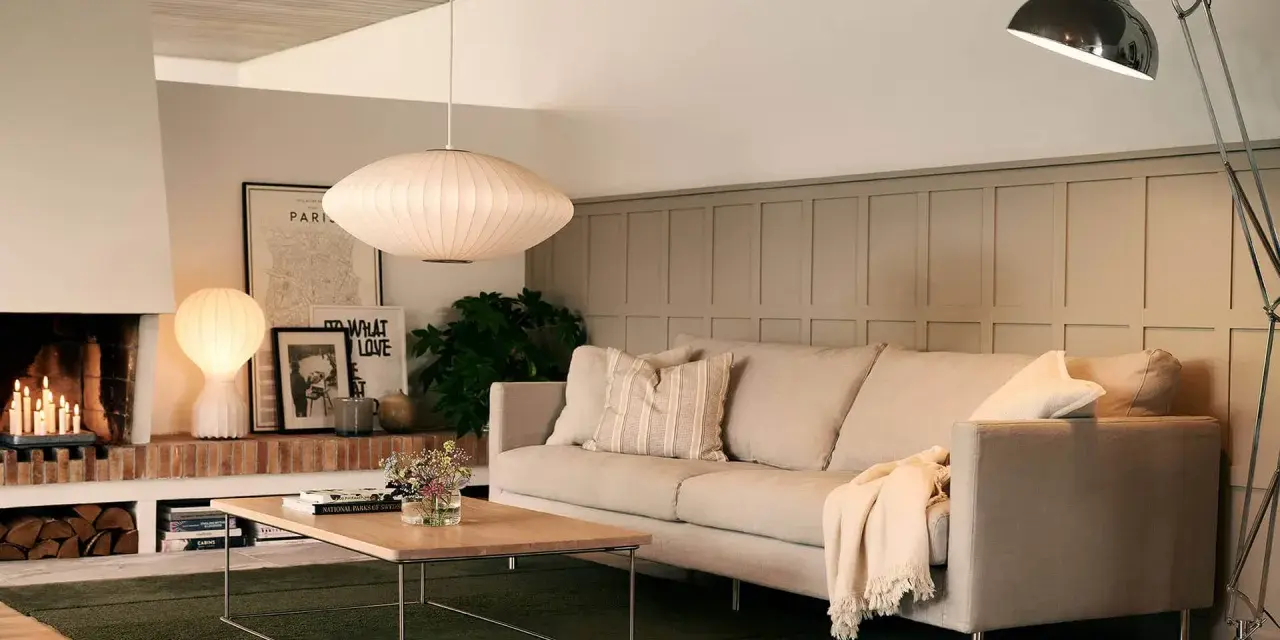 En ljus soffa med kuddar och en filt, framför ett soffbord i trä. En golvlampa och en taklampa lyser upp rummet. Perfekt för att mäta soffa mått.