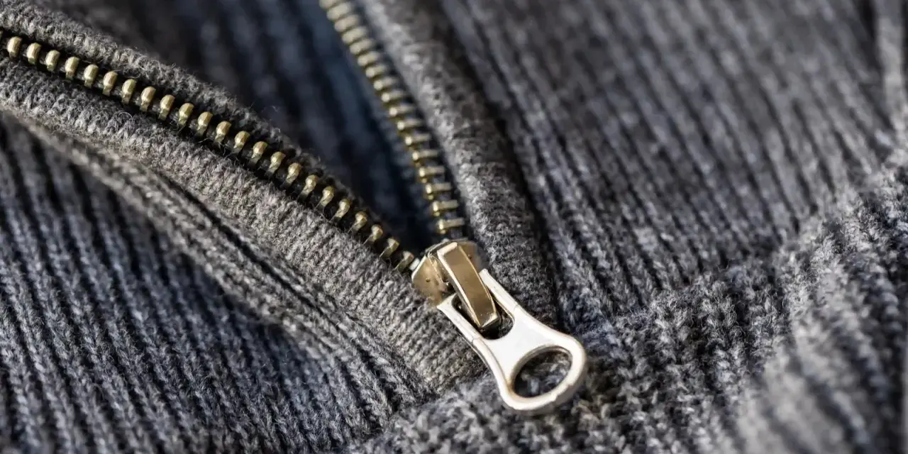 Metalowy suwak na szarym, dzianinowym materiale. Zamek jest częściowo rozpięty, co pokazuje, w którą stronę otwiera się zamek.