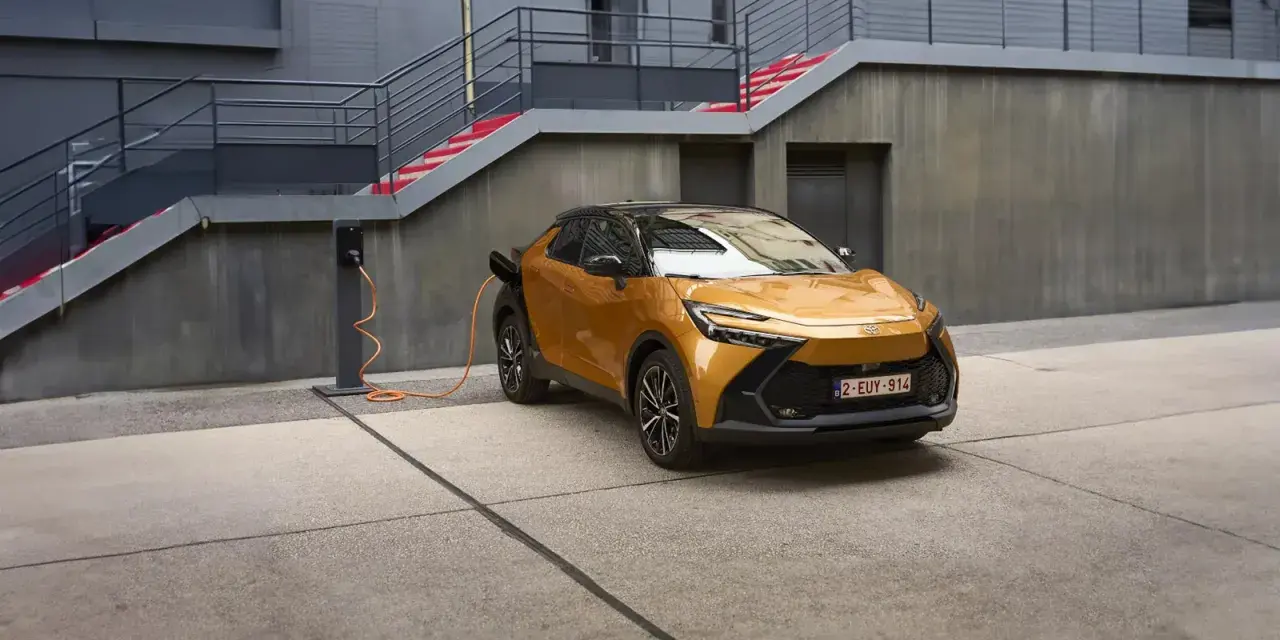 Złoty Toyota plug-in z pomarańczowym kablem ładowania podłączonym do stacji. Nowoczesna architektura w tle.