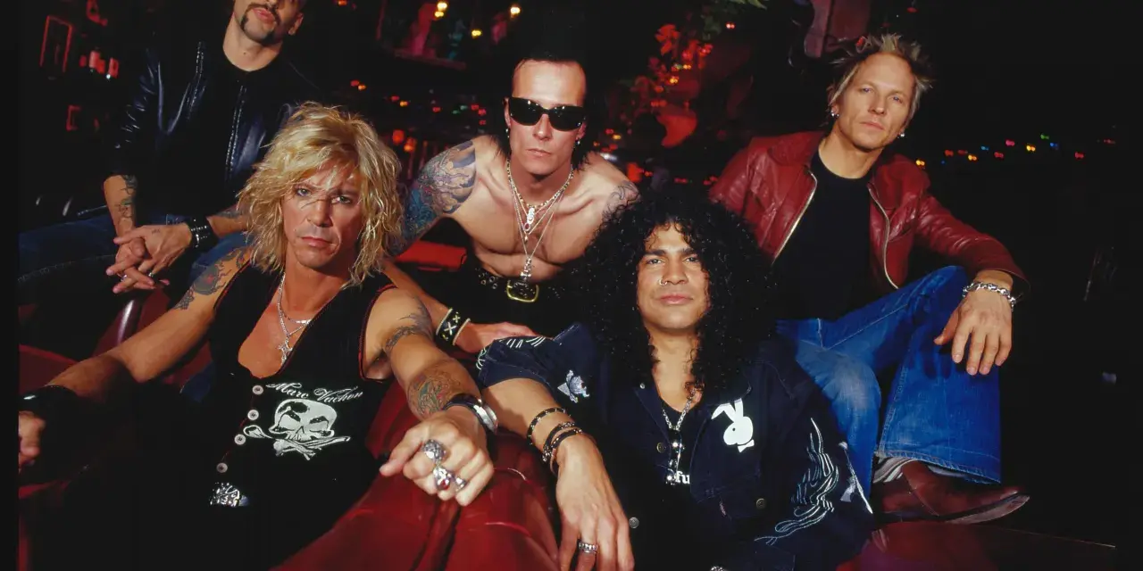 Cinq musiciens, un groupe de slash, posent sur un canapé rouge. Ils portent des vestes en cuir et des bijoux.