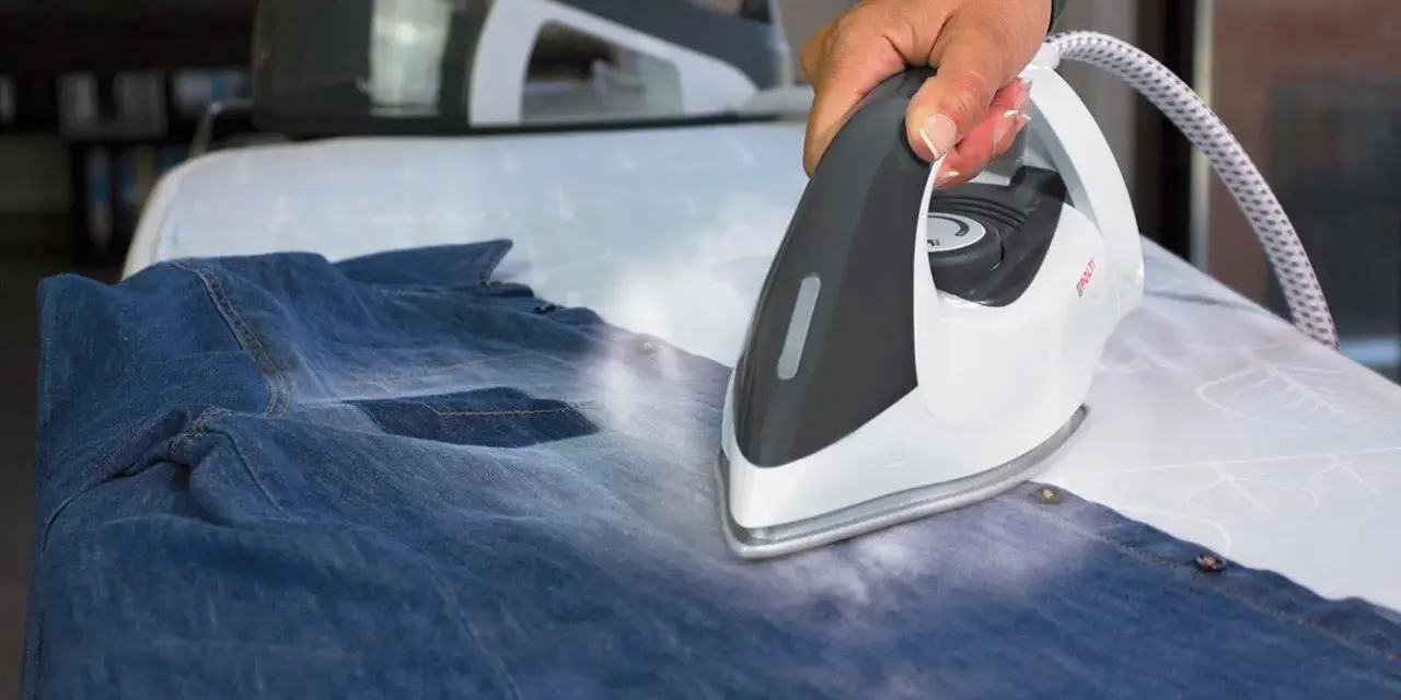 Hand using a steam iron on a denim shirt on an ironing board.