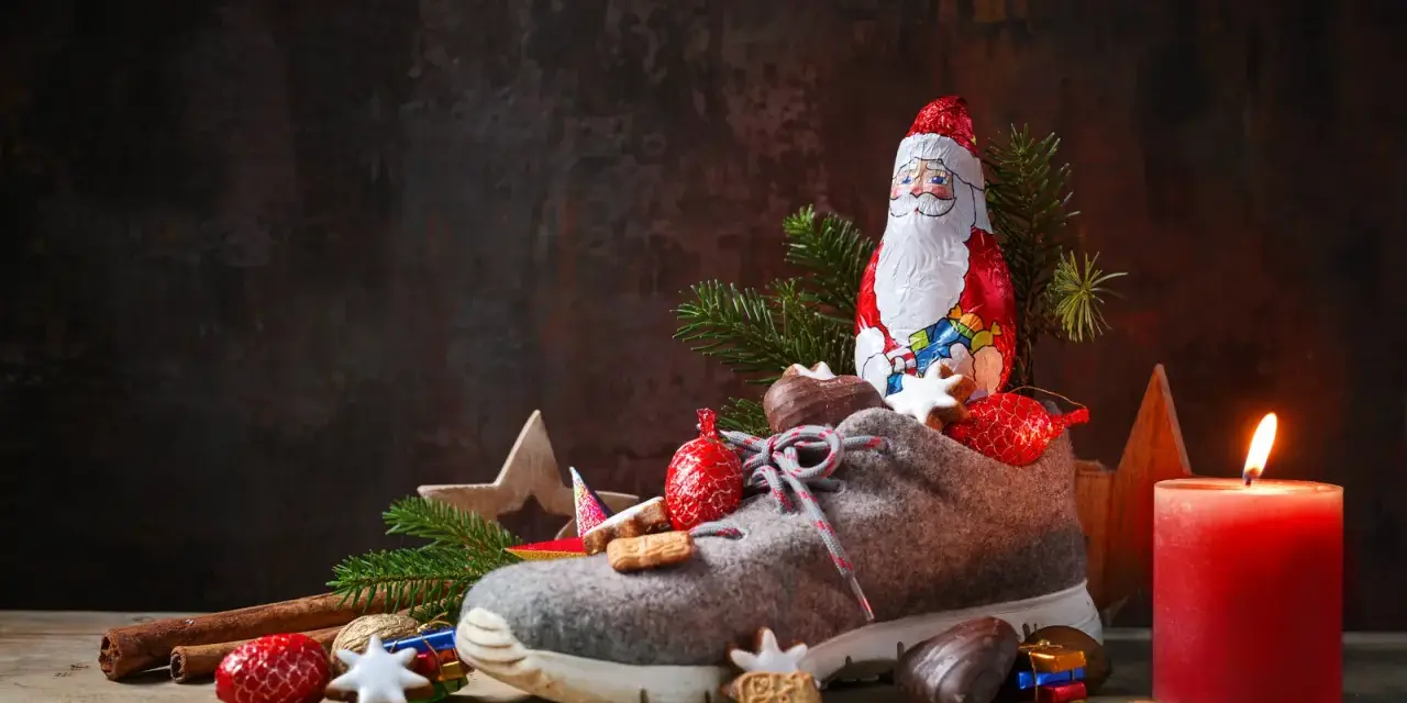 Ein Schuh gefüllt mit Leckereien, daneben ein Schokoladen-Nikolaus, Tannenzweige und eine brennende Kerze.