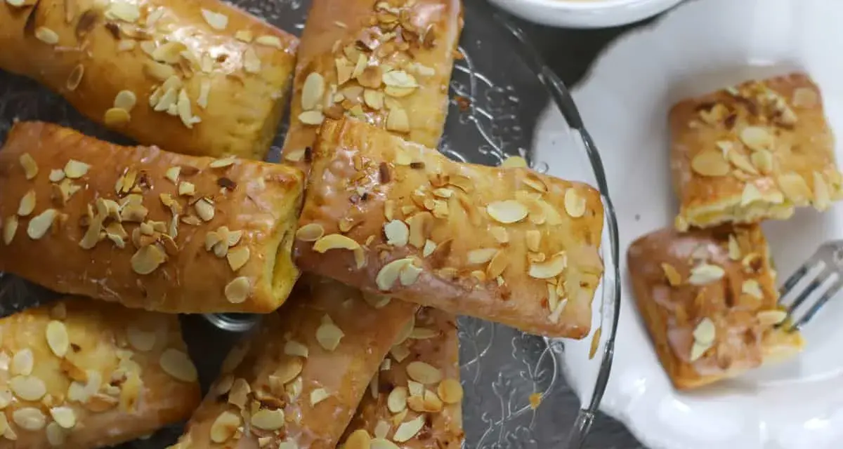Omas Apfeltaschen Rezept: goldbraune, glasierte Gebäckstücke mit Mandelblättchen, serviert auf einem Glasständer und einem Teller, daneben eine Tasse Kaffee.