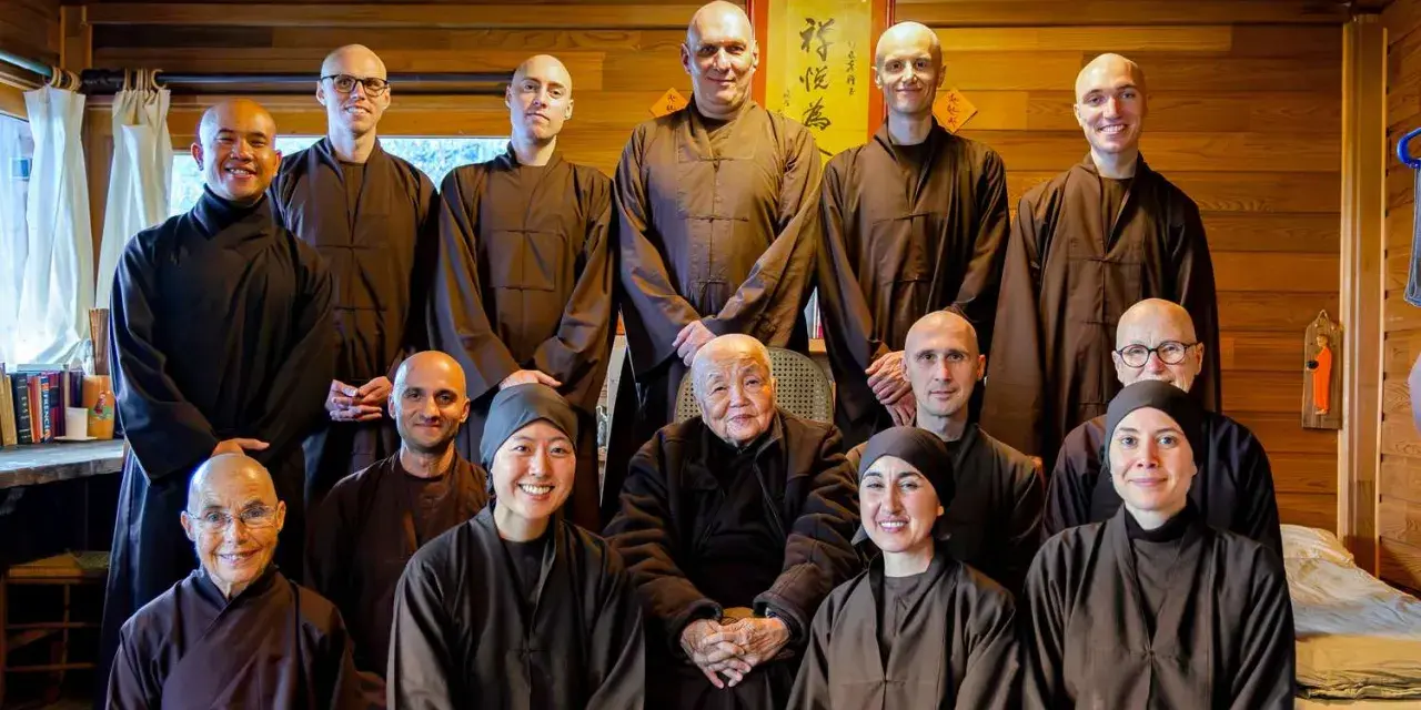 A group of monks and nuns, including an elder monk person in the center, pose for a photo in a room with wooden walls.