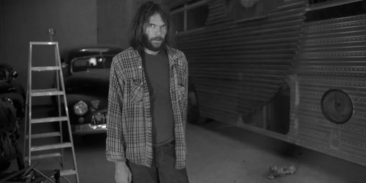 Neil Young, jung und nachdenklich, steht vor einem umgebauten Bus. Eine Leiter und ein Oldtimer im Hintergrund.