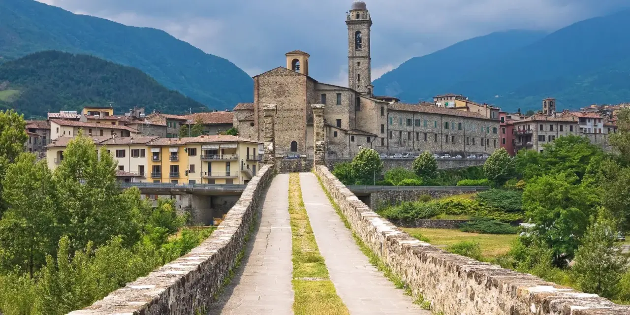Ponte medievale in pietra che conduce a un borgo con chiesa e campanile, incastonato tra le verdi montagne degli Appennini Emilia Romagna.