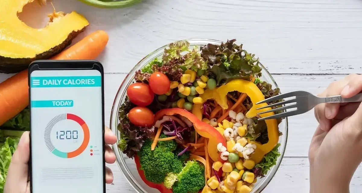 Aplikacja pokazuje 1200 kcal, podczas gdy ręka sięga widelcem po świeżą sałatkę.