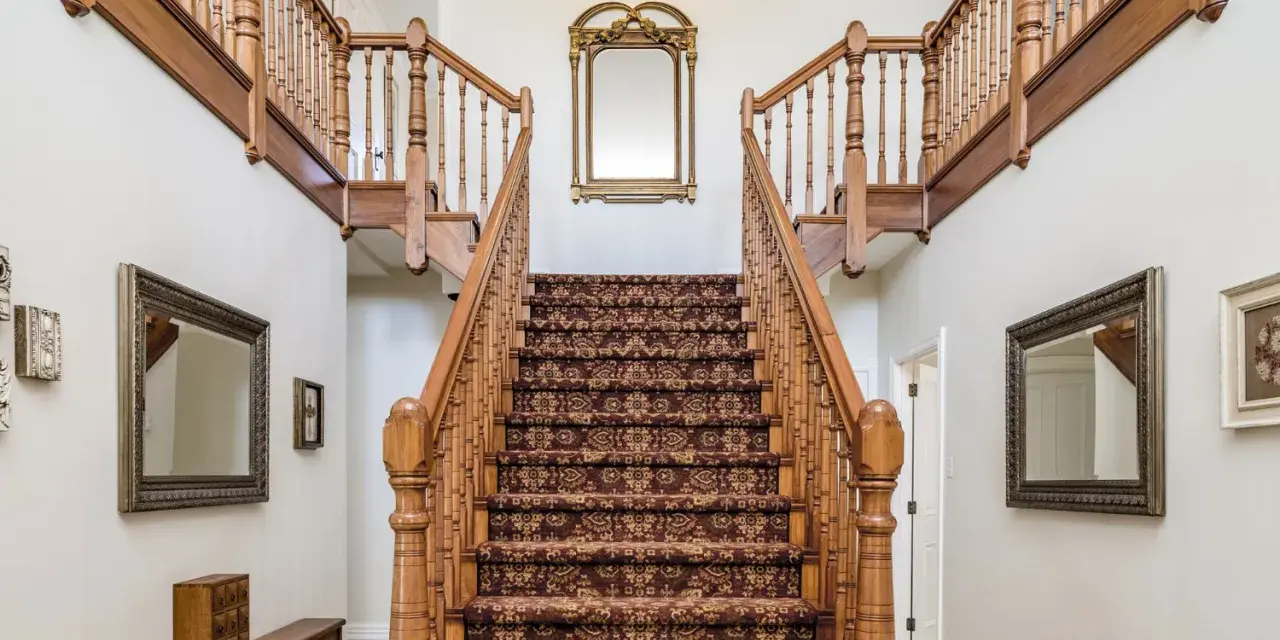 Elegante escalera antigua con alfombra de estilo persa y barandillas de madera. Un espejo dorado decora la pared central, complementando el revestir escalera antigua.