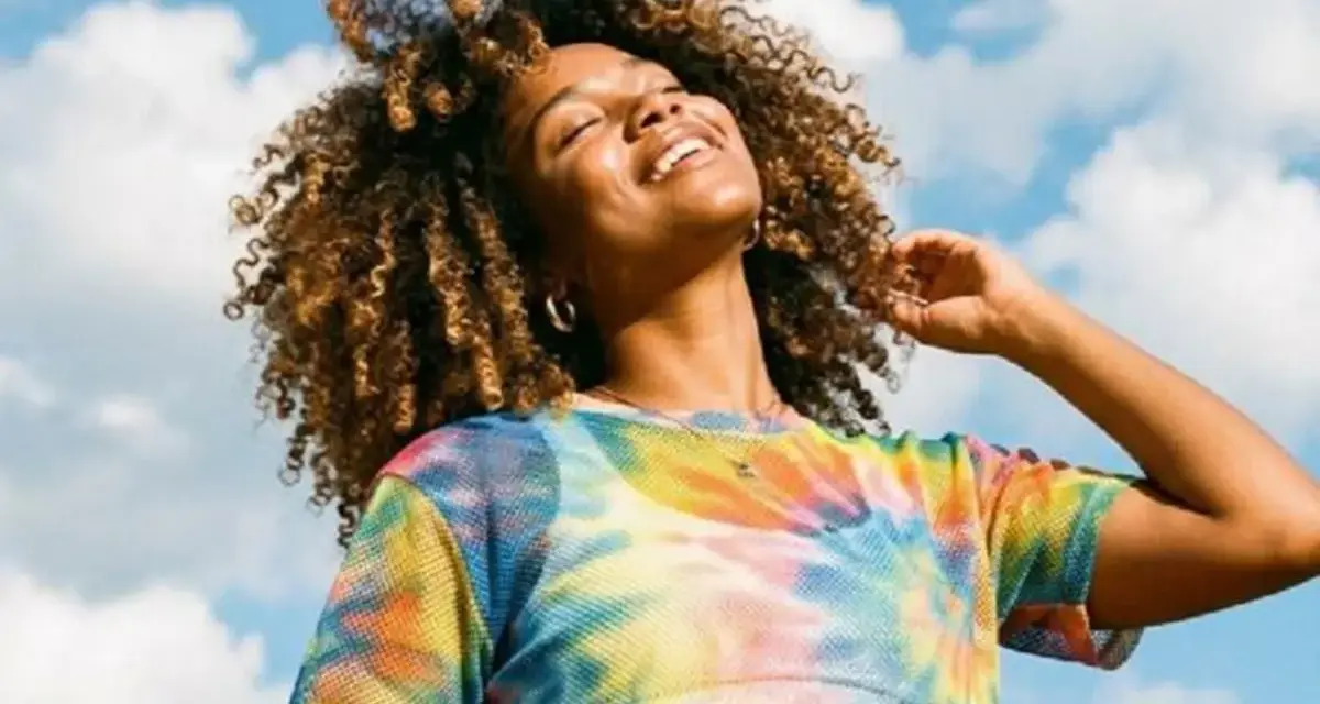 Une femme souriante aux cheveux bouclés, sous un ciel bleu nuageux. Elle porte un t-shirt tie-dye coloré. Une belle image pour apprendre comment rendre une photo flou.