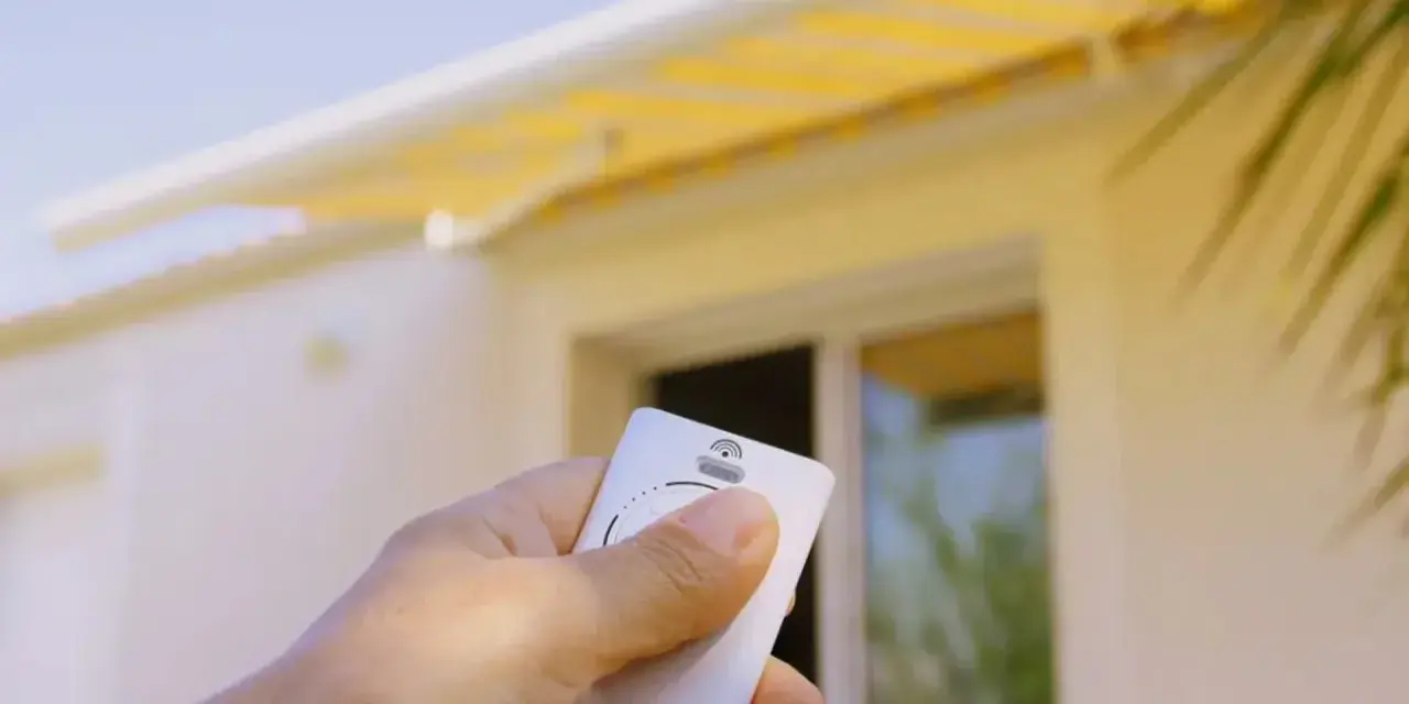 Main tenant une télécommande pour le réglage store banne manuel. Le store jaune est déployé sur une maison blanche.