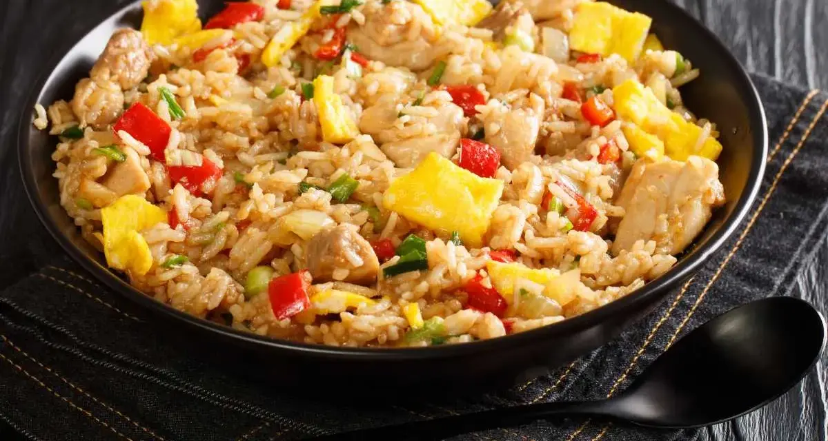 Un plato humeante de arroz chaufa con trozos de pollo, huevo revuelto, pimientos rojos y cebolla verde.