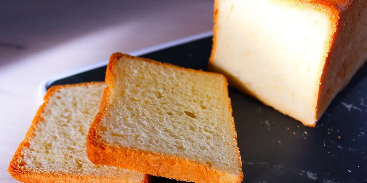 Pain de mie moelleux, parfait pour une recette pain de mie boulangerie maison. Deux tranches dorées et un pain entier sur un plateau sombre.