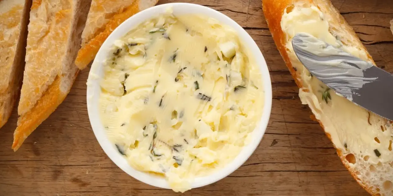 Knoblauchbutter selber machen ist einfach! Hier ist eine Schüssel mit selbstgemachter Knoblauchbutter, daneben Baguettescheiben und ein Messer, das Butter auf ein Brot streicht.