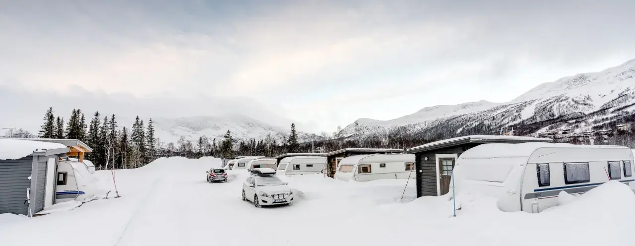 Vinterlandskap vid moskosel camping med snötäckta husvagnar och bilar, berg i bakgrunden.