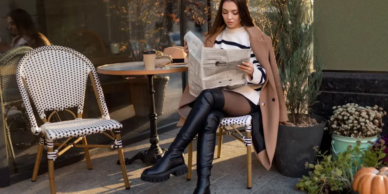 Eleganckie kozaki z szeroką cholewką idealnie dopełniają jesienną stylizację. Kobieta czyta gazetę przy stoliku w kawiarni.