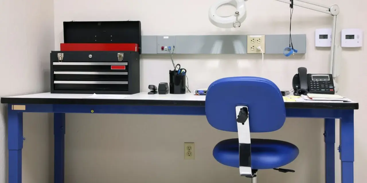 Warsztat z niebieskim stołem, na którym stoi skrzynka z narzędziami, telefon i lampa. Jaka wysokość stołu warsztatowego jest idealna do pracy?