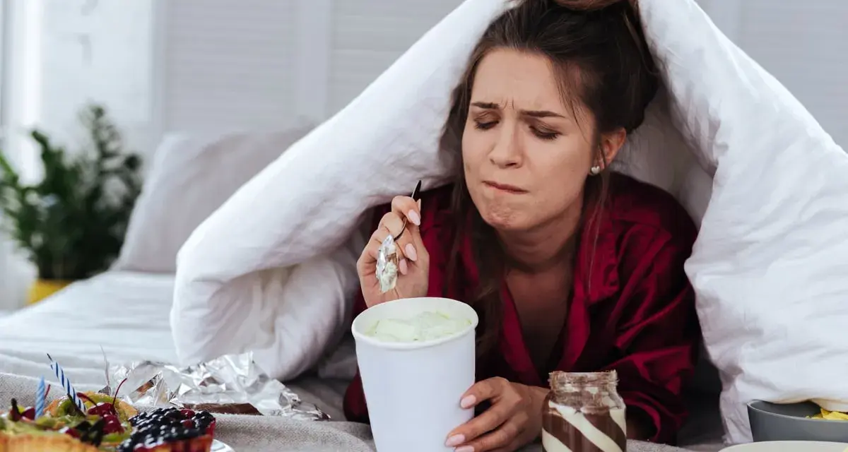 Ragazza sotto le coperte mangia gelato, dolci e pasta. La sua espressione suggerisce una forte fame pre ciclo.