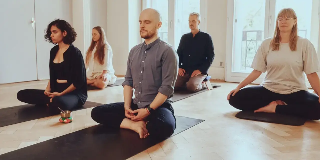 Menschen in einem hellen Raum meditieren auf Matten. Die Meditation wirkt beruhigend und fördert die Konzentration.