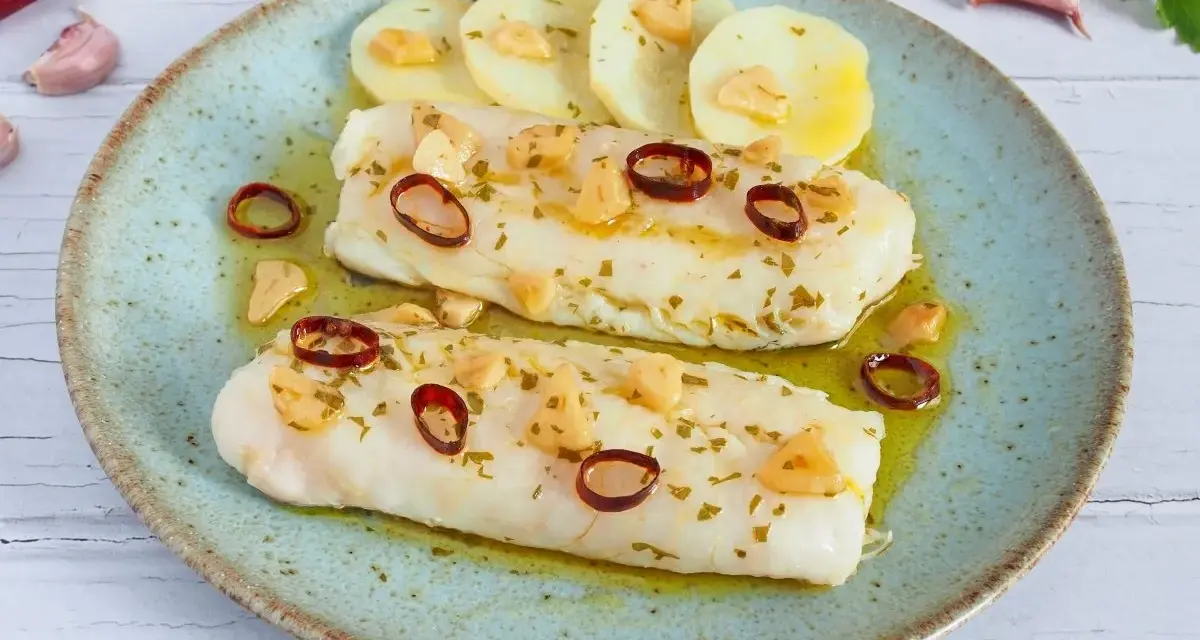 Dos filetes de merluza a la bilbaina, con patatas, ajo laminado y guindilla, en un plato azul.