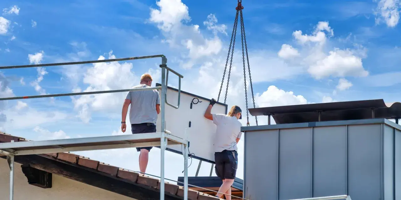 Dwaj pracownicy montują panel fotowoltaiczny na dachu. Ciężka praca i ryzyko wypadku to negatywne skutki fotowoltaiki, które często się pomija.