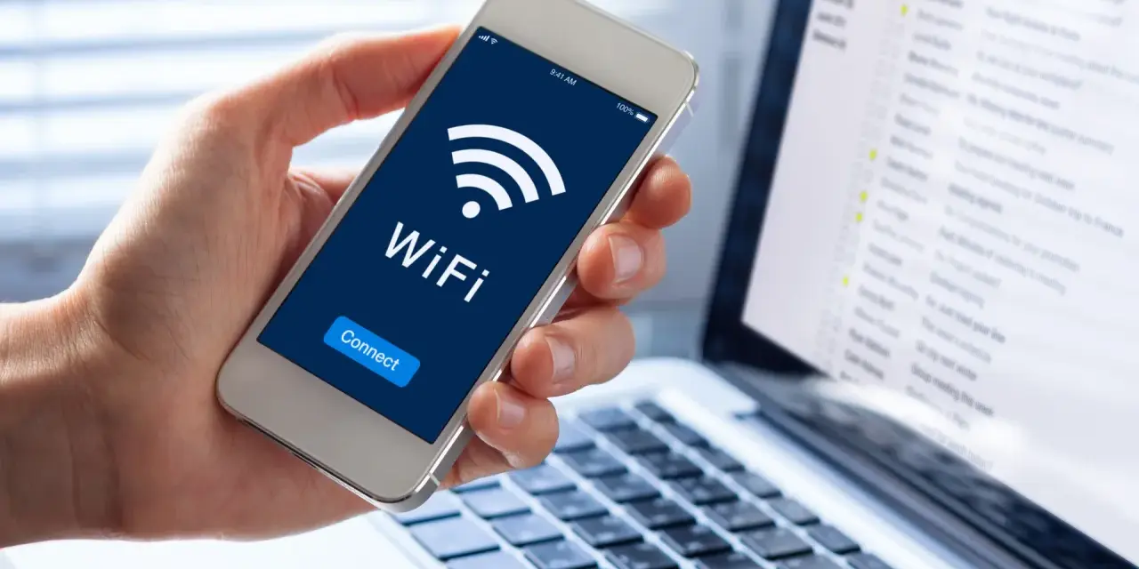 Dłoń trzyma smartfon z ikoną WiFi i napisem "Connect". Obok widać laptopa z listą e-maili, sugerującą pracę zdalną i dostęp do sieci, być może przez **ssid** domowy.