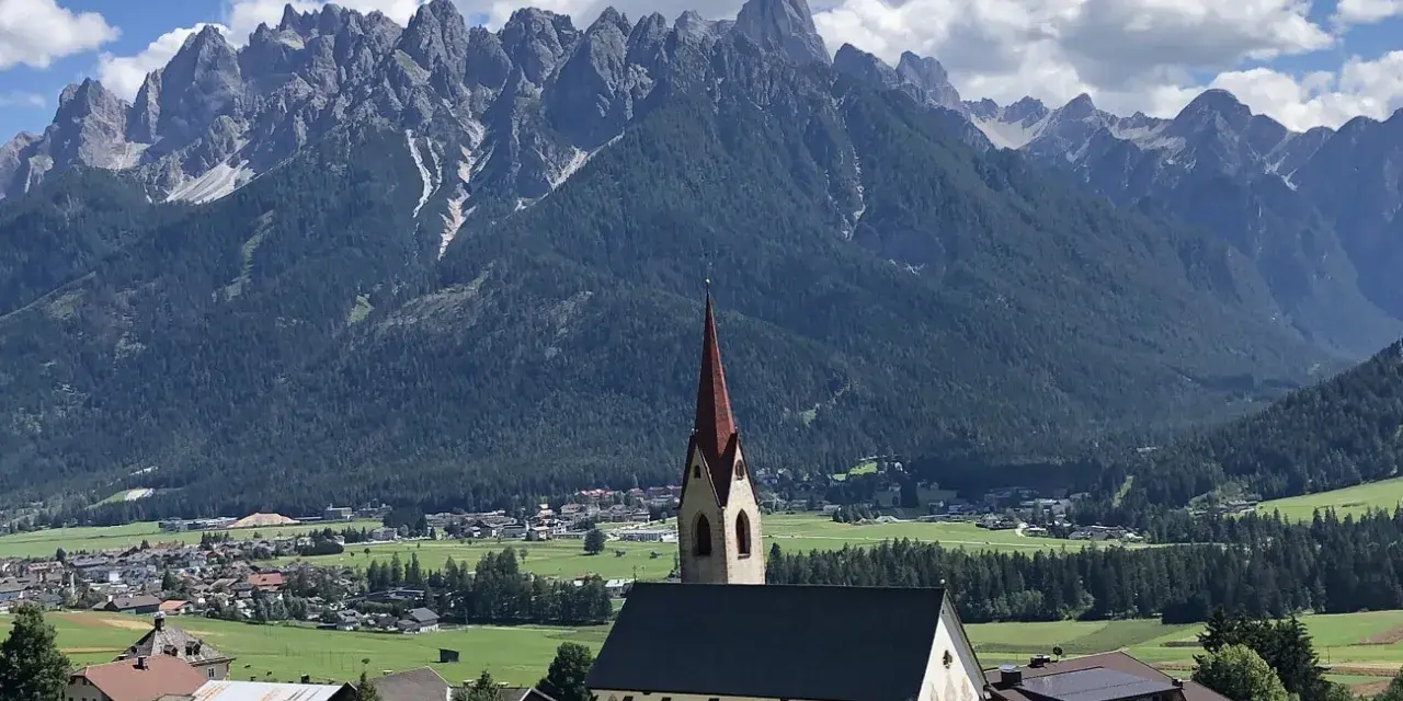 Chiesa con campanile rosso in primo piano, sullo sfondo le maestose vette delle Dolomiti e i **paesi più belli della Val Pusteria**.