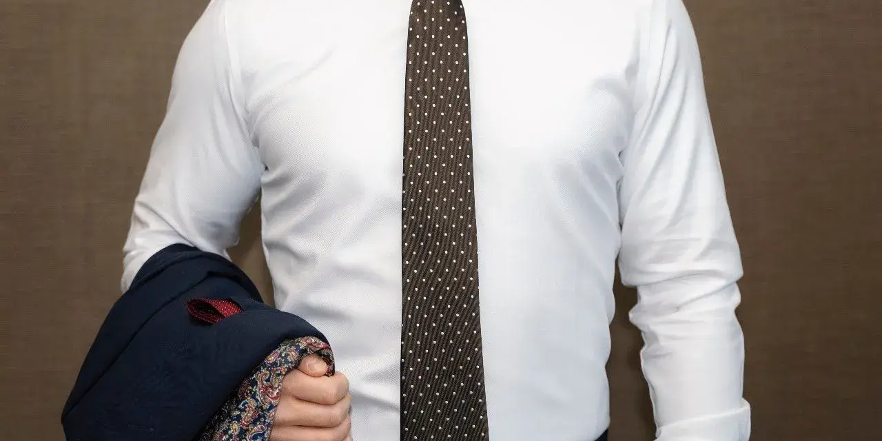 Elegancki mężczyzna w białej koszuli i modnym krawacie męskim w kropki, trzymający granatową marynarkę z wzorzystą podszewką.