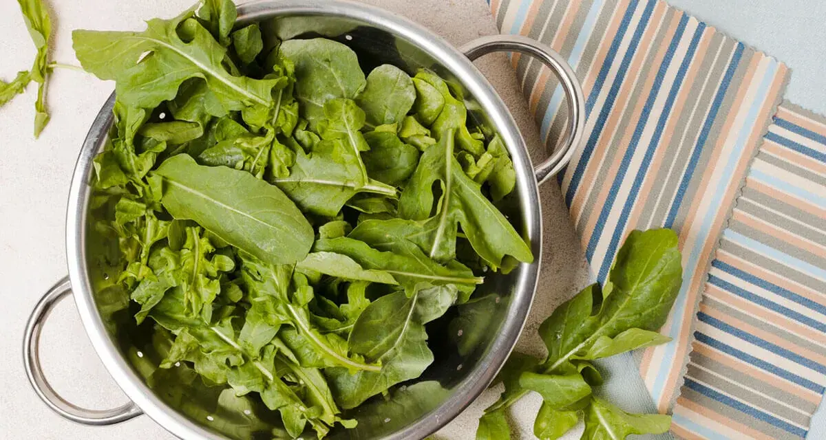 Rukola w durszlaku, idealna do przygotowania jadłospisu tygodniowego dieta FODMAP. Świeże liście na tle kolorowych tkanin.