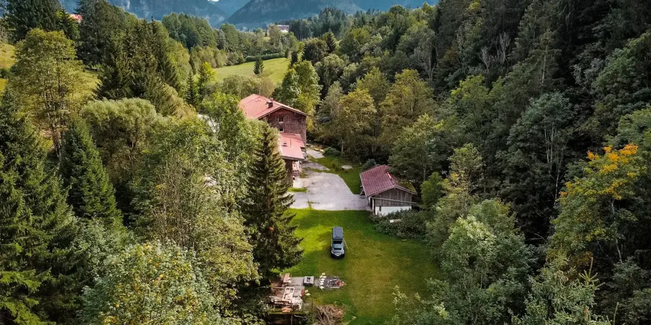 Idyllisches **Camping in den Bergen**: Ein Wohnmobil steht auf einer grünen Wiese neben einem Bach, umgeben von dichten Wäldern und traditionellen Holzhäusern.