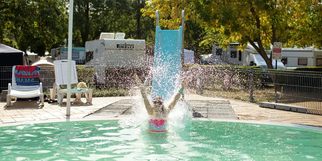 Radość na aranypart camping! Dziecko zjeżdża ze zjeżdżalni prosto do basenu, tworząc fontannę wody.