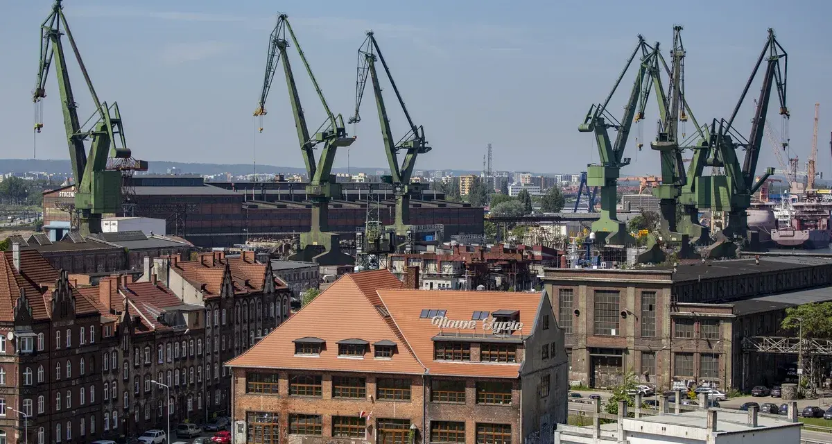Widok na stocznię w Gdańsku z charakterystycznymi dźwigami. W oddali budynek z napisem "Nowe Syrie", nawiązujący do muzeum sztuki współczesnej Gdańsk.