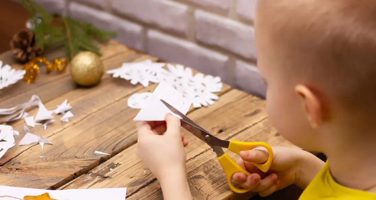 Małe rączki wycinają śnieżynkę z papieru. Na stole leżą gotowe ozdoby i gałązka choinki.