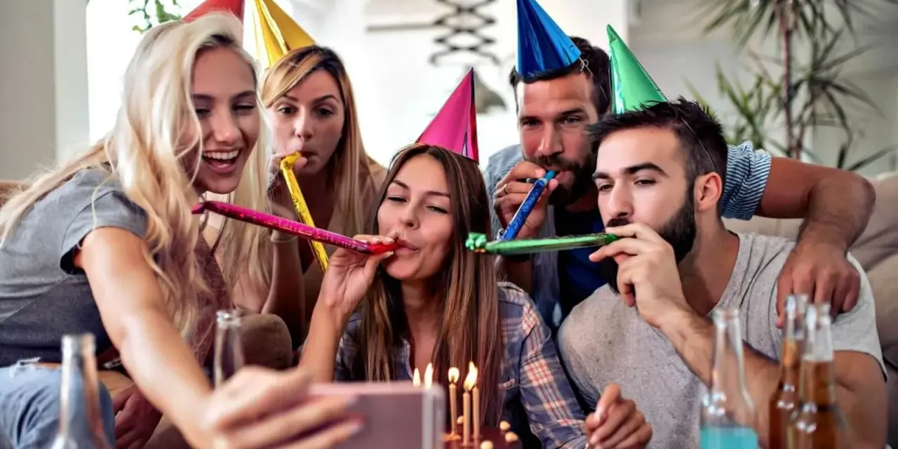 Radosne urodziny przyjaciółki! Grupa przyjaciół w kolorowych czapkach świętuje, dmucha w trąbki i robi selfie przy torcie.