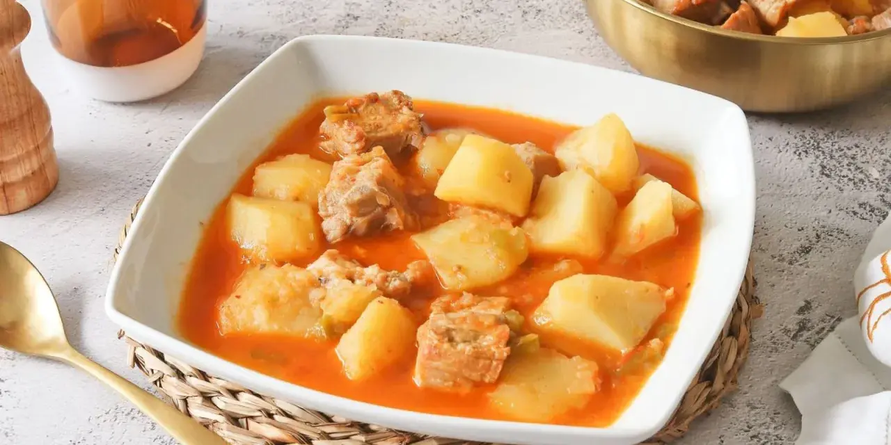 Un reconfortante plato de patatas con costillas en una salsa rojiza, servido en un cuenco blanco.