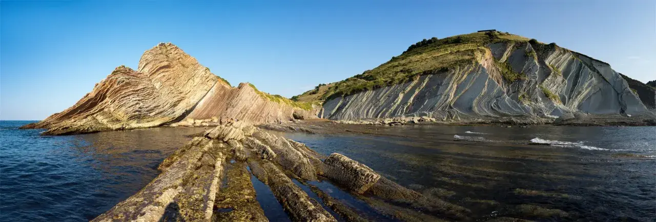 The flysch de Zumaia showcases dramatic, layered rock formations meeting the sea under a clear blue sky.