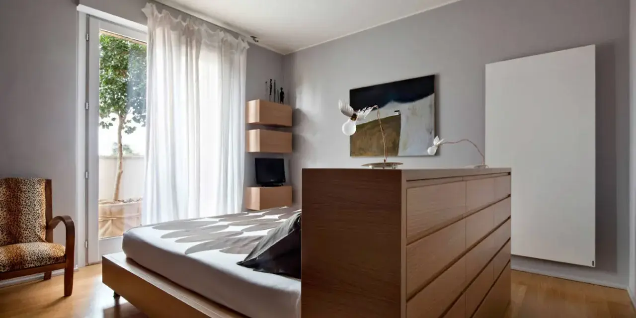Un moderno **letto in mezzo alla stanza**, con un copriletto a motivi floreali, accanto a una poltrona leopardata e una cassettiera in legno.