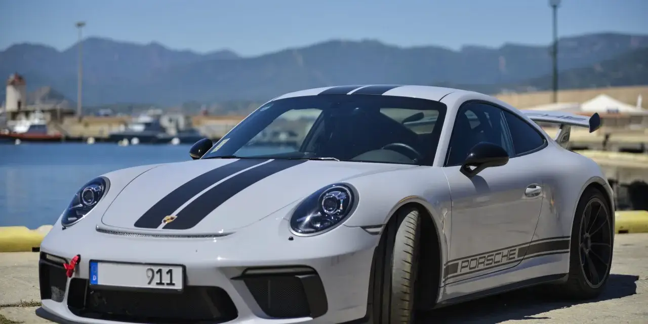Szary Porsche 911 z czarnymi pasami, stojący nad wodą. Zastanawiasz się nad porsche 911 cena?