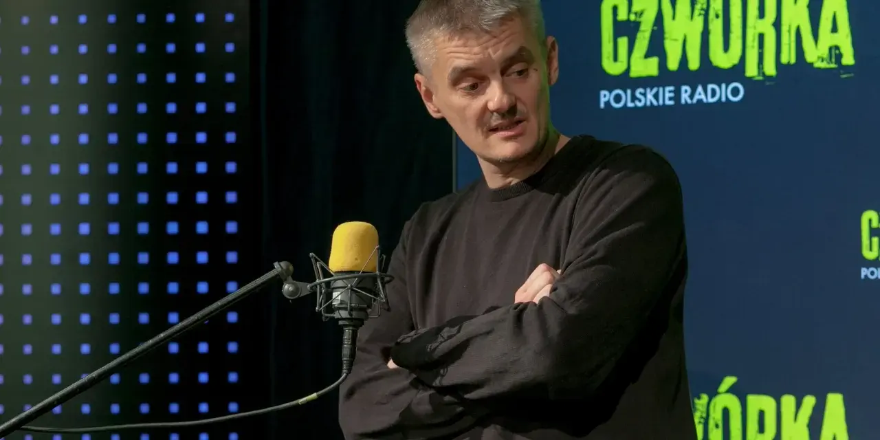 Jacek Sienkiewicz w studiu Polskiego Radia, przed mikrofonem, z rękami skrzyżowanymi na piersi.