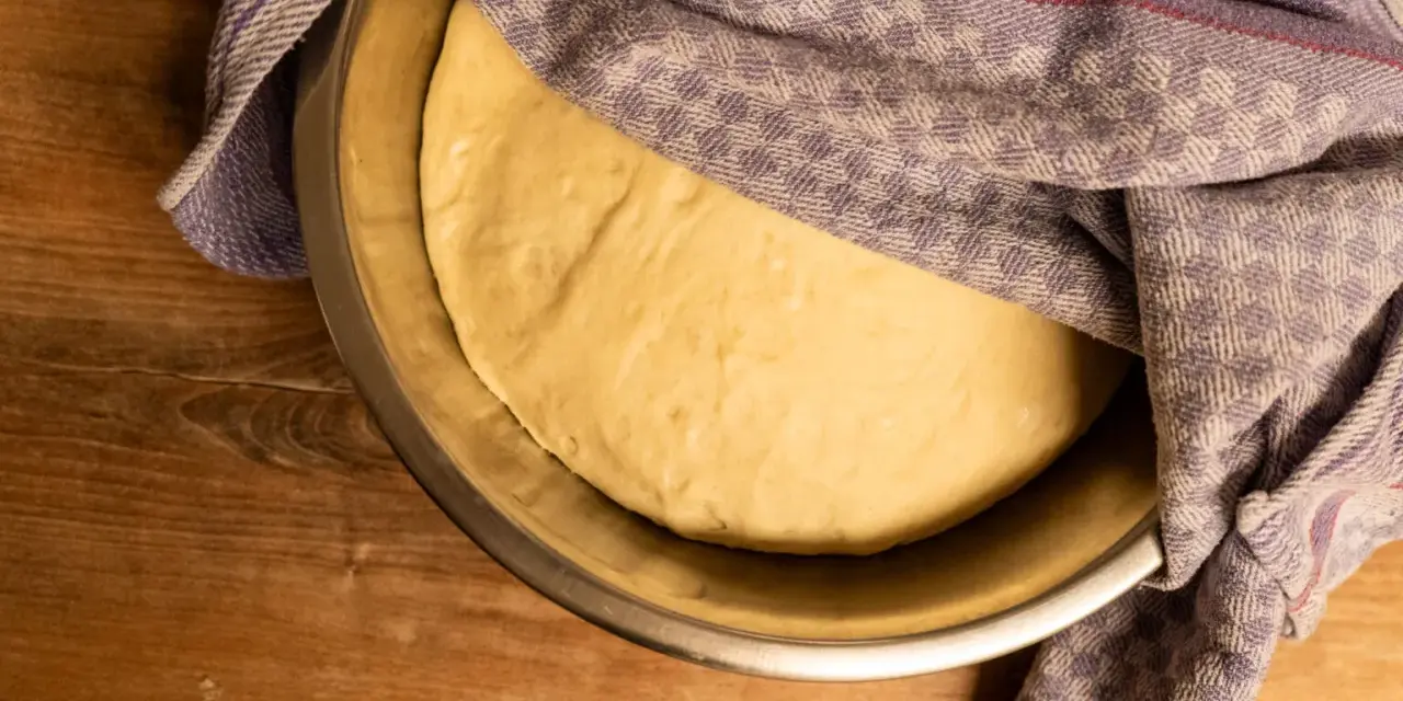 Ein geschmeidiger Teig ruht in einer Schüssel, bedeckt mit einem karierten Tuch, bereit für das Backen.