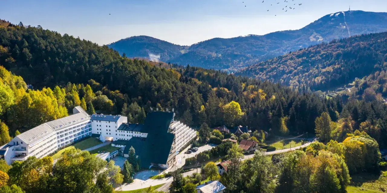 Malowniczy hotel przy stoku narciarskim w Polsce, otoczony jesiennym lasem. W oddali góry i niebo z ptakami.