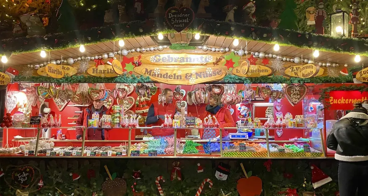 A vibrant Dresden Christmas market stall overflowing with gingerbread hearts, candied nuts, and colorful sweets. Festive lights twinkle above the tempting displays.
