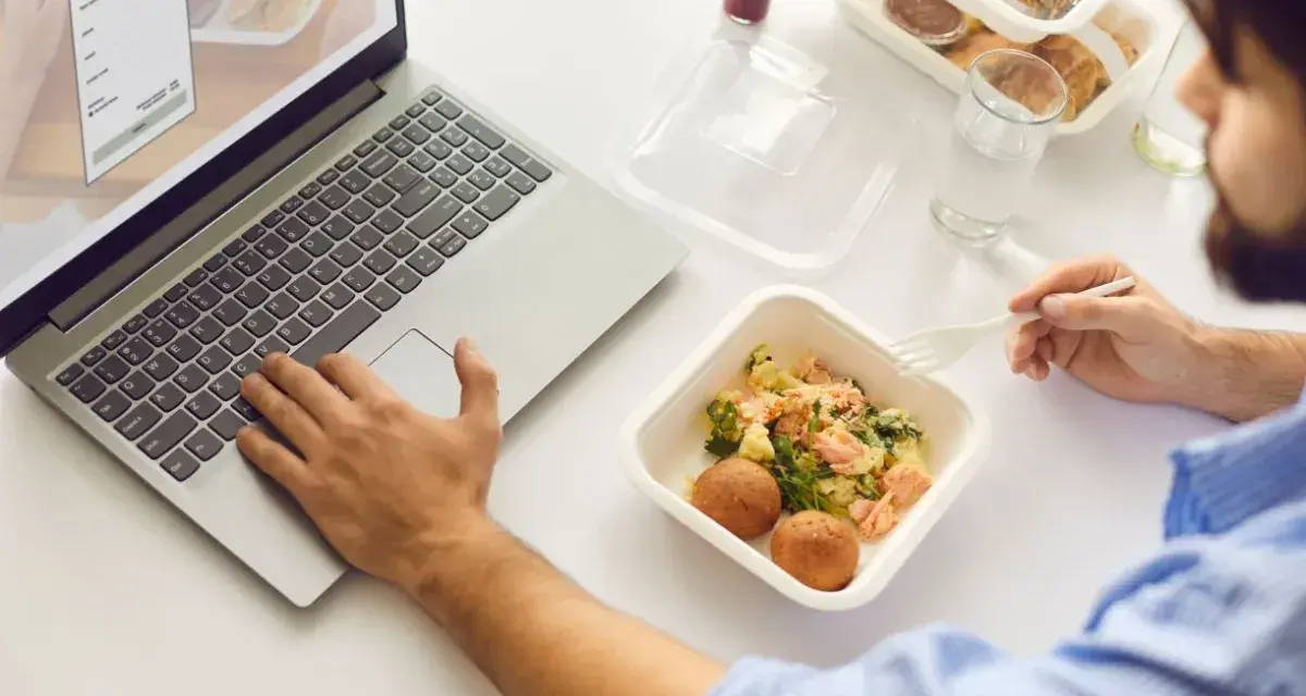 Mężczyzna je dietetyczny lunch z pudełka, pracując na laptopie.