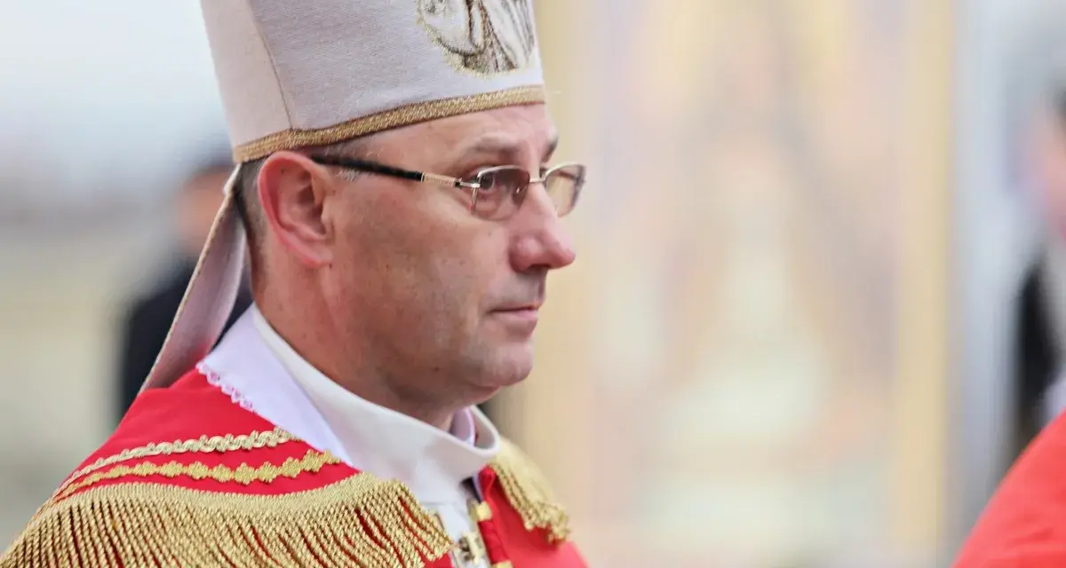 Biskup w stroju pontyfikalnym, z mitrą na głowie, w okularach. W tle widać fragment obrazu.