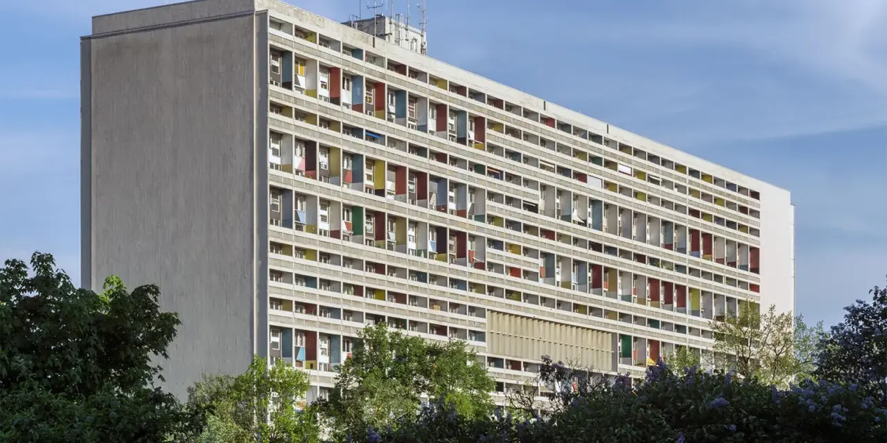 Długi budynek mieszkalny w stylu modernizm, z rzędami balkonów w kolorowych obramowaniach, otoczony zielenią.