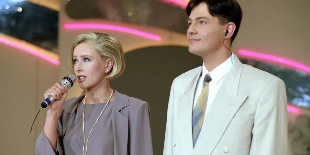 Kto prowadził czar par? Ona z mikrofonem, on w jasnej marynarce, na tle neonowych świateł.