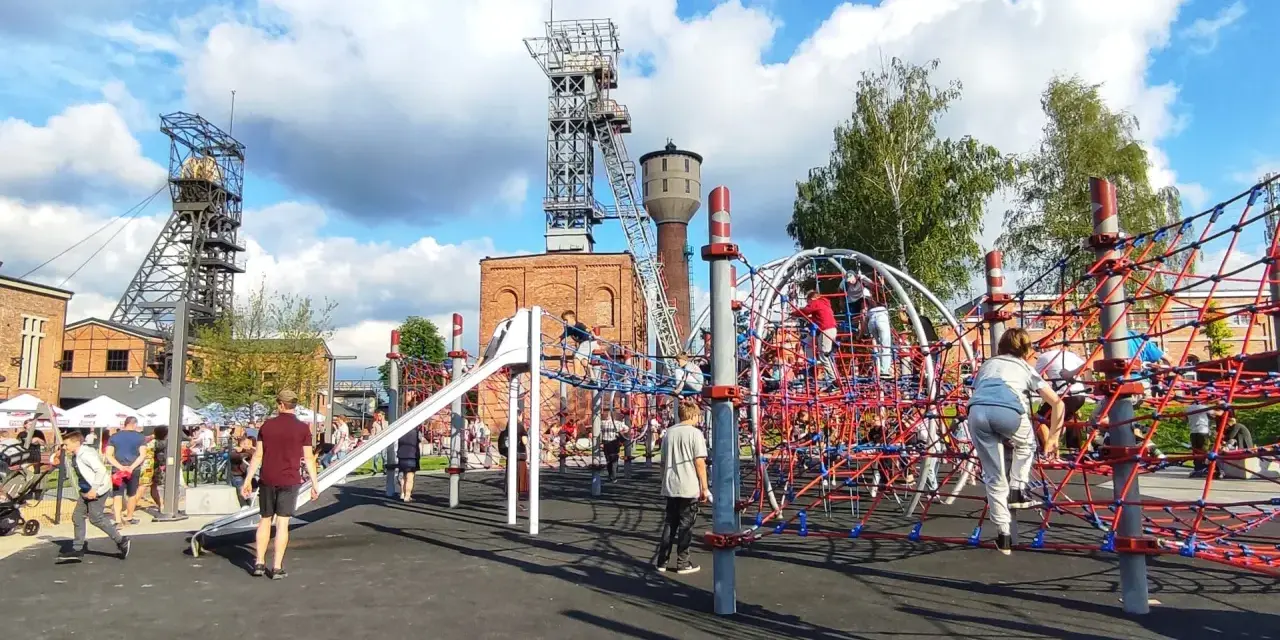Rybnik atrakcje: plac zabaw dla dzieci z linowymi konstrukcjami i zjeżdżalnią, w tle wieże szybowe kopalni.