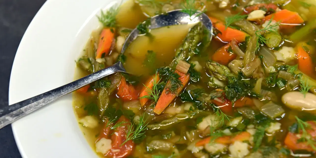 Un fumante minestrone di verdure, ricco di carote, fagioli e erbe aromatiche, servito in una ciotola bianca con un cucchiaio.