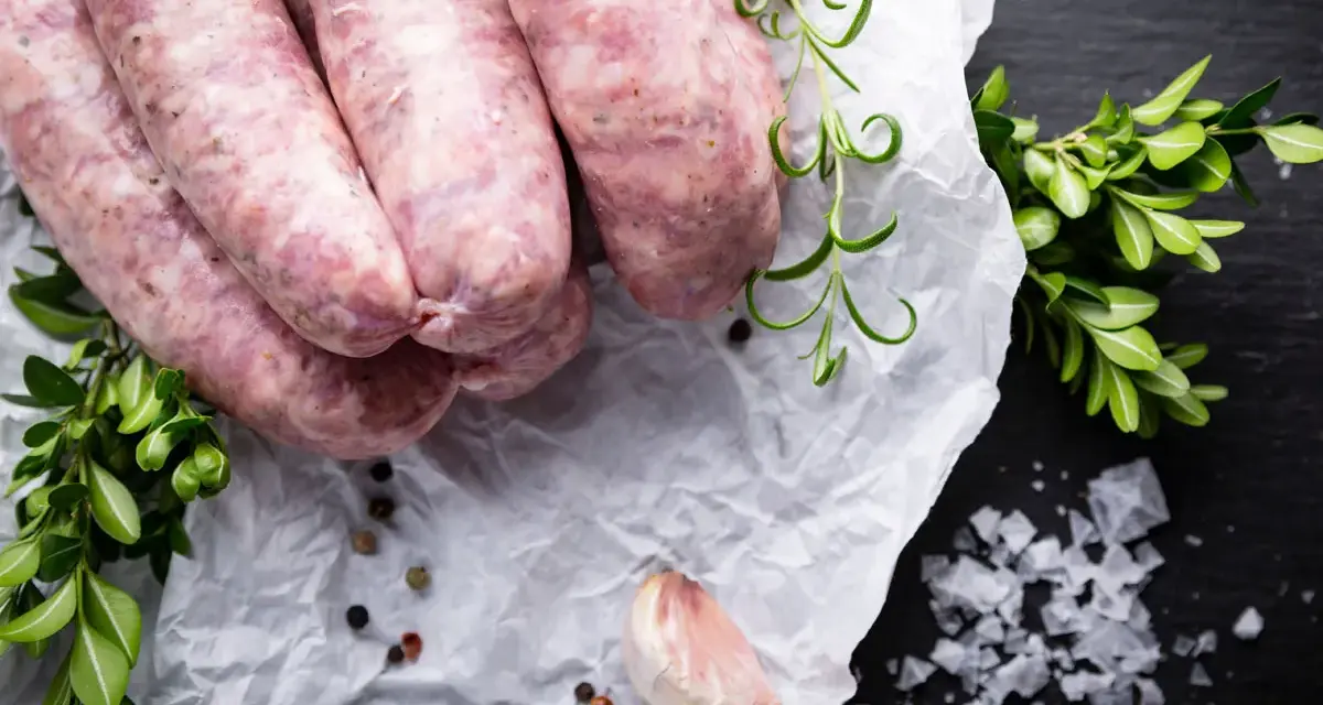 Surowa biała kiełbasa na papierze, obok gałązki rozmarynu i czosnek. Zastanawiasz się nad biała kiełbasa kcal?
