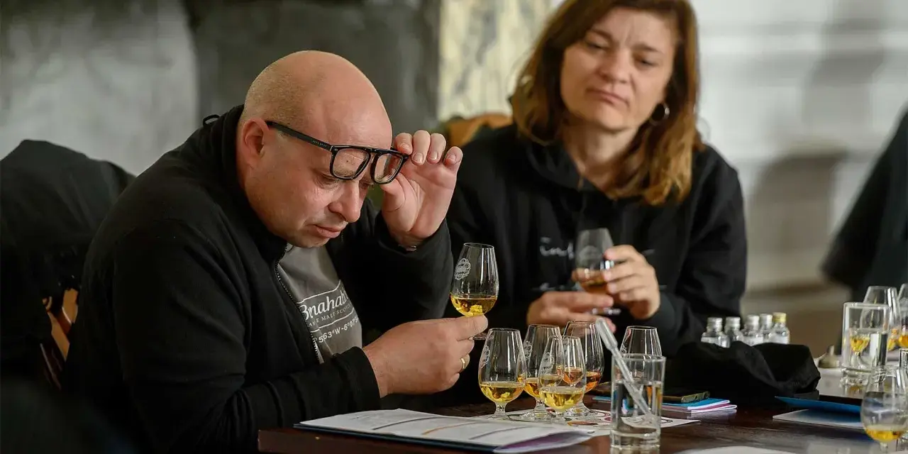Mann mit Brille prüft ein Glas mit bernsteinfarbenem blended malt whisky. Eine Frau im Hintergrund hält ebenfalls ein Glas.
