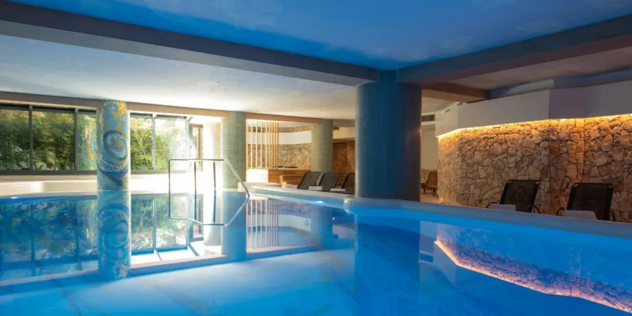 Indoor swimming pool with lounge chairs and a stone wall with warm lighting.
